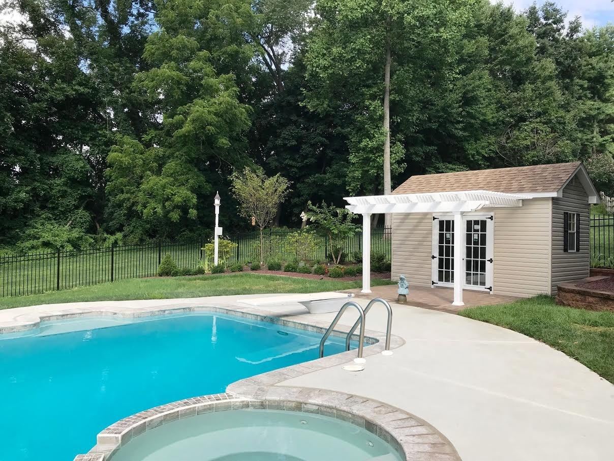 Inground gunite pool with a brick-coped spa, white pergola and pool house framed by mature trees, built by JHL Pools near West Chester, PA.