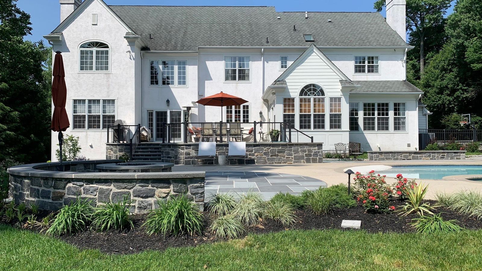 Backyard of a white Main Line Colonial home with stone-walled patio, bluestone hardscape, perennial gardens and an inground pool by JHL Pools in Chester County, PA.