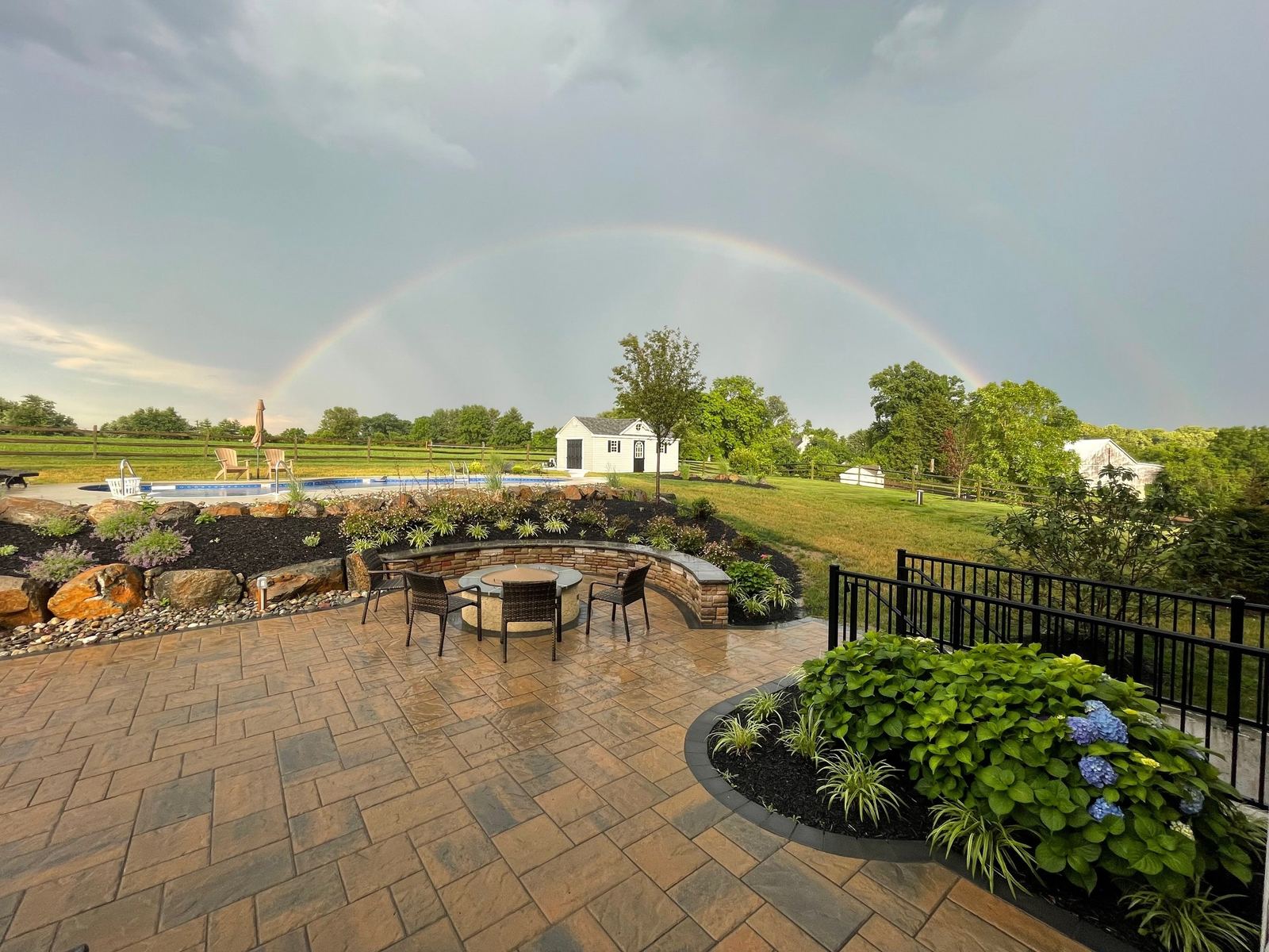 Paver patio with a circular stone fire-pit seating area overlooking an inground pool and lawn, a backyard resort project by JHL Pools in Chester County, PA.
